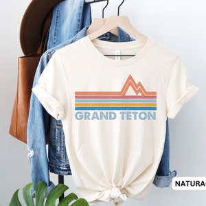 Puede incluir: Una camiseta de color crema con un dise&ntilde;o de monta&ntilde;a retro y el texto "GRAND TETON" en azul. La camiseta est&aacute; en una percha y tiene un color natural.