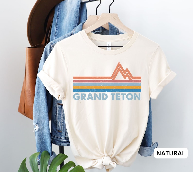 Puede incluir: Una camiseta de color crema con un dise&ntilde;o de monta&ntilde;a retro y el texto "GRAND TETON" en azul. La camiseta est&aacute; en una percha y tiene un color natural.