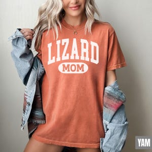 May include: Orange short-sleeved t-shirt with the text "LIZARD MOM" in white block letters. The word "MOM" is in a white oval. The shirt is worn with a blue denim jacket.