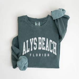 May include: A teal-colored sweatshirt with the text "ALYS BEACH FLORIDA" in white block letters. The sweatshirt is folded, with the sleeves rolled up. The fabric appears soft and comfortable.