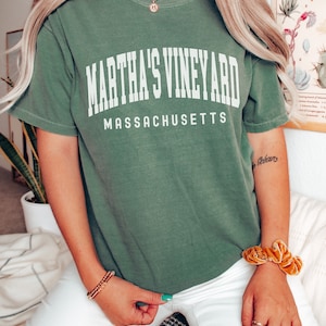May include: A green t-shirt with the text "Martha's Vineyard Massachusetts" printed on the front. The shirt is worn by a person with long blonde hair and white pants.