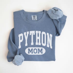 Könnte beinhalten: Ein staubblaues Sweatshirt mit dem weißen Schriftzug "PYTHON MOM". Der Text ist über einem Oval mit der Aufschrift "MOM" angeordnet. Die Ärmel sind umgeschlagen, und das Sweatshirt liegt flach.