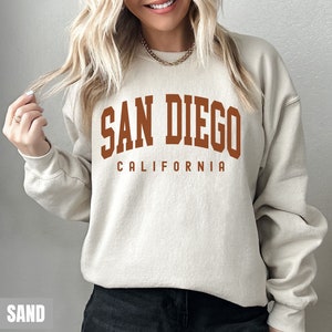 Pode incluir: Uma camisa de moletom bege com uma estampa gr&aacute;fica marrom que diz "San Diego California".