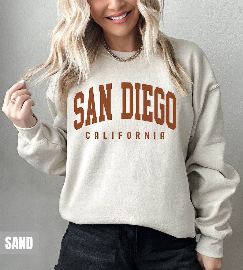 Pode incluir: Uma camisa de moletom bege com uma estampa gr&aacute;fica marrom que diz "San Diego California".