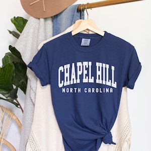Op de afbeelding: Een marineblauw t-shirt met witte tekst die "Chapel Hill North Carolina" zegt.