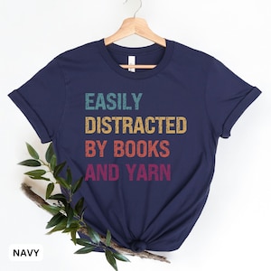 May include: A navy blue t-shirt with the text "EASILY DISTRACTED BY BOOKS AND YARN" in a vintage rainbow color scheme.