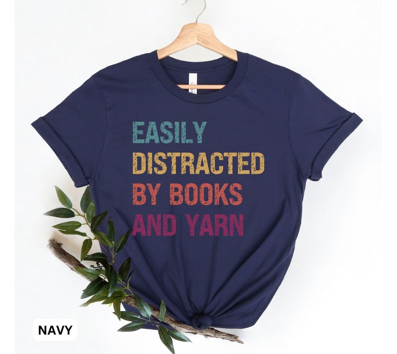 May include: A navy blue t-shirt with the text "EASILY DISTRACTED BY BOOKS AND YARN" in a vintage rainbow color scheme.