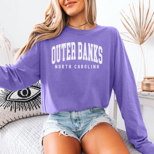 May include: A long-sleeved purple sweatshirt with the text "Outer Banks North Carolina" in white. The sweatshirt is paired with denim shorts. The image is taken indoors.