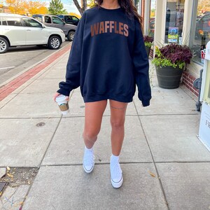 May include: A black sweatshirt with the word "WAFFLES" printed in brown letters across the chest. The sweatshirt is being worn by a person with white sneakers.