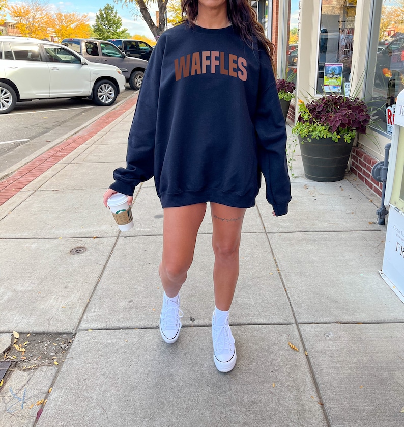 May include: A black sweatshirt with the word "WAFFLES" printed in brown letters across the chest. The sweatshirt is being worn by a person with white sneakers.