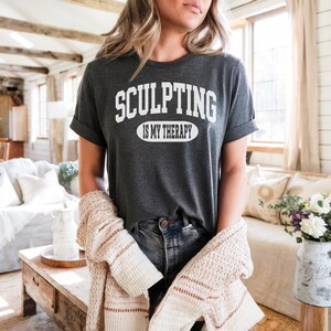 K&ouml;nnte beinhalten: Dunkelgraues T-Shirt mit dem wei&szlig;en Text "SCULPTING IS MY THERAPY". Das Shirt hat hochgekrempelte &Auml;rmel. Ein beigefarbener Cardigan ist &uuml;ber den Armen drapiert. Das Model tr&auml;gt eine blaue Jeans.