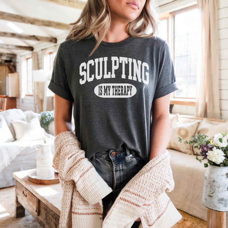 K&ouml;nnte beinhalten: Dunkelgraues T-Shirt mit dem wei&szlig;en Text "SCULPTING IS MY THERAPY". Das Shirt hat hochgekrempelte &Auml;rmel. Ein beigefarbener Cardigan ist &uuml;ber den Armen drapiert. Das Model tr&auml;gt eine blaue Jeans.