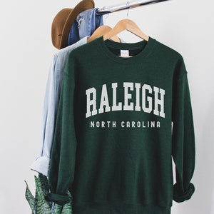 Pode incluir: Uma camisa de moletom verde escura com texto branco que diz "RALEIGH NORTH CAROLINA".