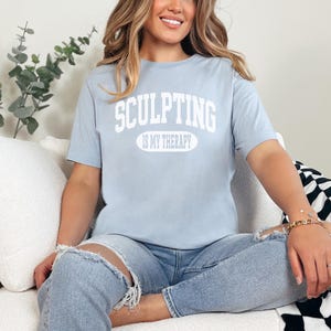 K&ouml;nnte beinhalten: Hellblaues T-Shirt mit dem wei&szlig;en Text "SCULPTING IS MY THERAPY". Die Person tr&auml;gt hellblaue Jeans mit zerrissenen Knien. Der Hintergrund umfasst ein wei&szlig;es Sofa und eine schwarz-wei&szlig; karierte Decke.