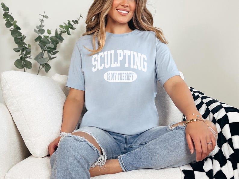 K&ouml;nnte beinhalten: Hellblaues T-Shirt mit dem wei&szlig;en Text "SCULPTING IS MY THERAPY". Die Person tr&auml;gt hellblaue Jeans mit zerrissenen Knien. Der Hintergrund umfasst ein wei&szlig;es Sofa und eine schwarz-wei&szlig; karierte Decke.