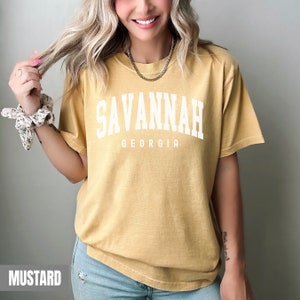 Op de afbeelding: Mosterdkleurig T-shirt met de tekst "SAVANNAH GEORGIA" in witte blokletters. Het shirt heeft korte mouwen en een relaxte pasvorm. Een zilveren ketting is zichtbaar.
