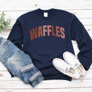 May include: A navy blue sweatshirt with the word "WAFFLES" printed in brown, pink, and orange letters. The sweatshirt is laid flat on a white wooden surface with a pair of white sneakers and blue jeans.