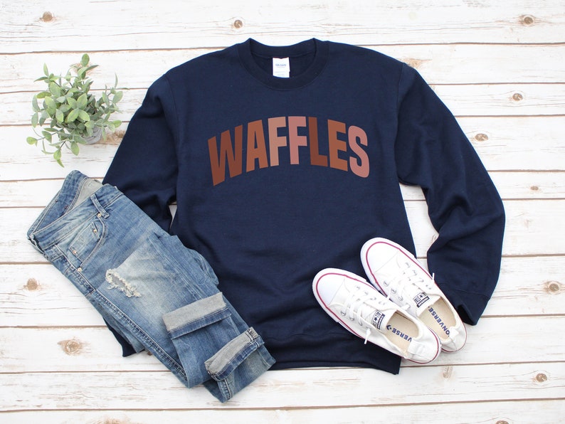 May include: A navy blue sweatshirt with the word "WAFFLES" printed in brown, pink, and orange letters. The sweatshirt is laid flat on a white wooden surface with a pair of white sneakers and blue jeans.