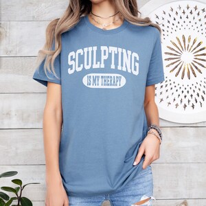 K&ouml;nnte beinhalten: Hellblaues T-Shirt mit dem wei&szlig;en Schriftzug "SCULPTING IS MY THERAPY". Das Shirt hat kurze &Auml;rmel und wird mit einer blauen Jeans getragen. Der Text ist in Gro&szlig;buchstaben gehalten.