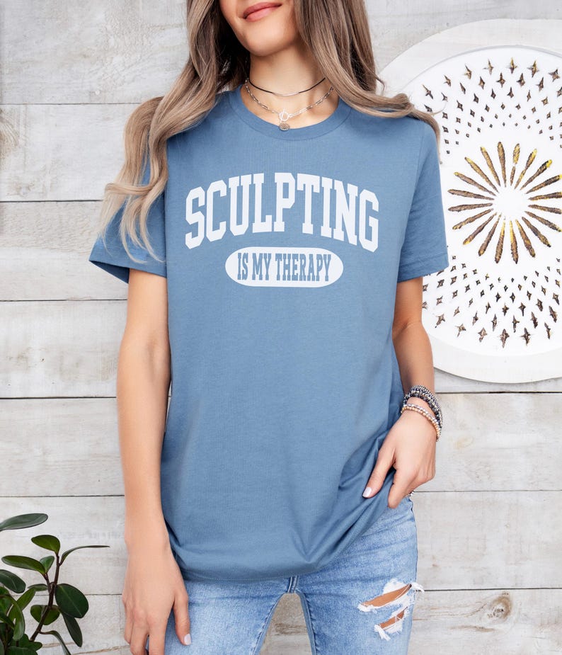 K&ouml;nnte beinhalten: Hellblaues T-Shirt mit dem wei&szlig;en Schriftzug "SCULPTING IS MY THERAPY". Das Shirt hat kurze &Auml;rmel und wird mit einer blauen Jeans getragen. Der Text ist in Gro&szlig;buchstaben gehalten.