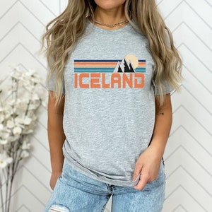 Puede incluir: Una camiseta gris con un gr&aacute;fico retro de monta&ntilde;as y una puesta de sol. El texto "ICELAND" est&aacute; impreso en naranja debajo del gr&aacute;fico.