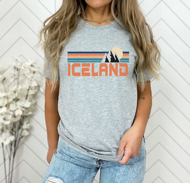Puede incluir: Una camiseta gris con un gr&aacute;fico retro de monta&ntilde;as y una puesta de sol. El texto "ICELAND" est&aacute; impreso en naranja debajo del gr&aacute;fico.