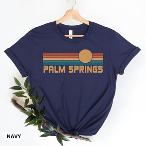 May include: Navy blue t-shirt with a retro-style graphic of a sunset over Palm Springs. The graphic features a yellow sun and multi-colored stripes.