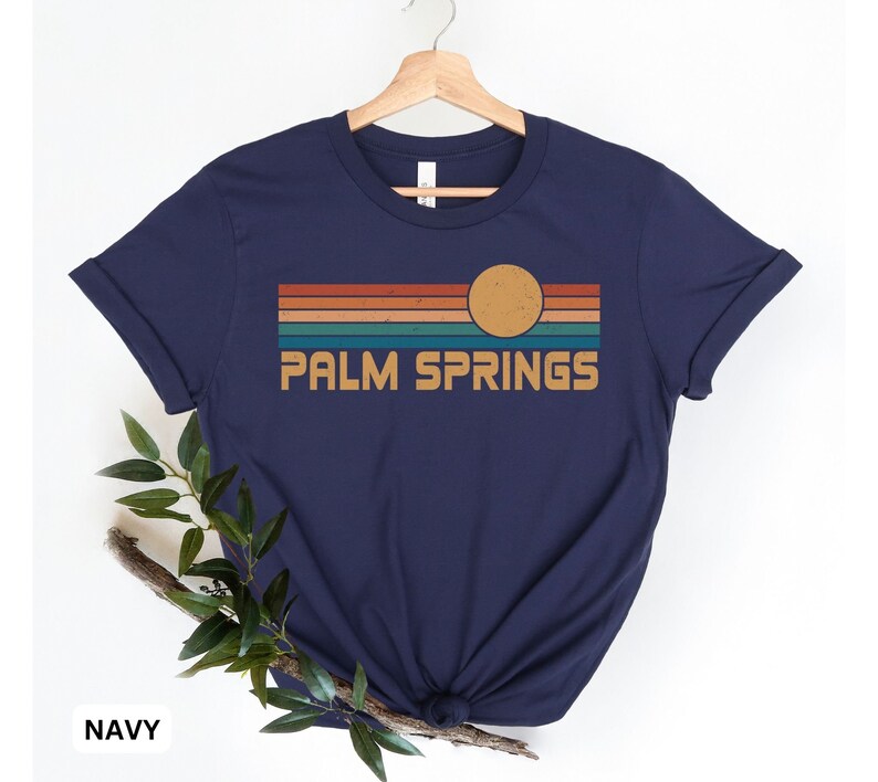 May include: Navy blue t-shirt with a retro-style graphic of a sunset over Palm Springs. The graphic features a yellow sun and multi-colored stripes.