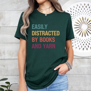 May include: A dark green t-shirt with the text "EASILY DISTRACTED BY BOOKS AND YARN" in a vintage style font with colorful accents.