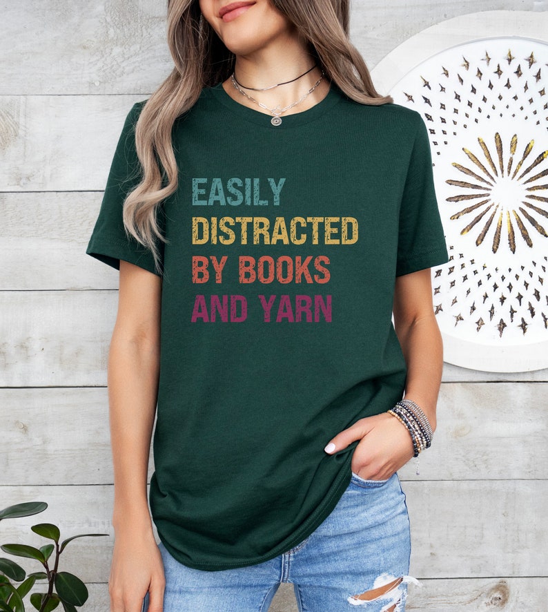 May include: A dark green t-shirt with the text "EASILY DISTRACTED BY BOOKS AND YARN" in a vintage style font with colorful accents.