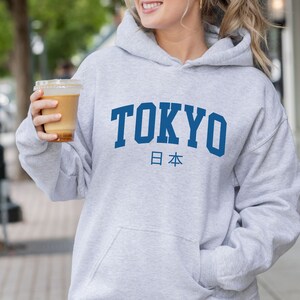 以下が含まれることがあります： グレーのフード付きスウェットシャツに、青い文字で「TOKYO」と、その下に青い文字で日本の日本語「日本」が書かれています。