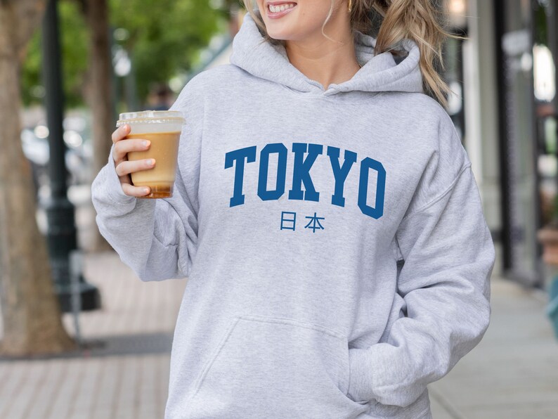 以下が含まれることがあります： グレーのフード付きスウェットシャツに、青い文字で「TOKYO」と、その下に青い文字で日本の日本語「日本」が書かれています。