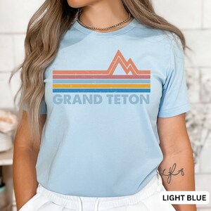 Puede incluir: Camiseta azul claro con un gr&aacute;fico retro de monta&ntilde;as y rayas en tonos naranja, amarillo, azul y gris. El texto "GRAND TETON" est&aacute; impreso debajo del gr&aacute;fico.