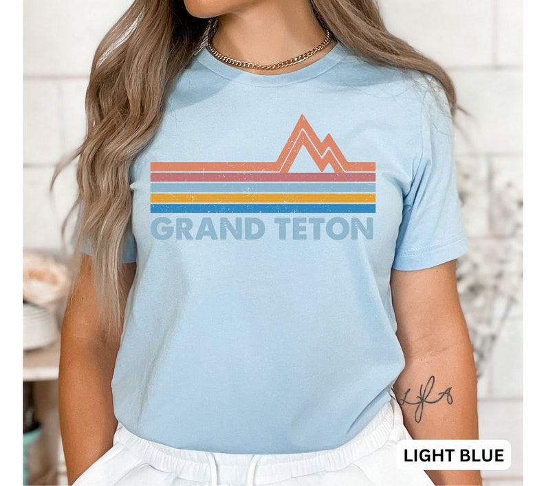 Puede incluir: Camiseta azul claro con un gr&aacute;fico retro de monta&ntilde;as y rayas en tonos naranja, amarillo, azul y gris. El texto "GRAND TETON" est&aacute; impreso debajo del gr&aacute;fico.