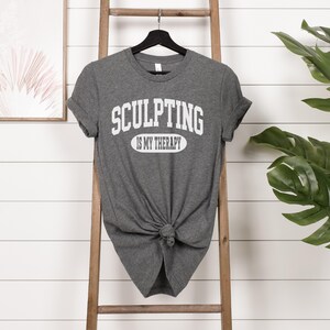 K&ouml;nnte beinhalten: Ein graues T-Shirt mit dem Text "SCULPTING IS MY THERAPY" in wei&szlig;en Blockbuchstaben. Das Shirt ist an der Taille geknotet und h&auml;ngt an einem schwarzen Kleiderb&uuml;gel. Eine Holzleiter und eine gr&uuml;ne Pflanze sind im Hintergrund.
