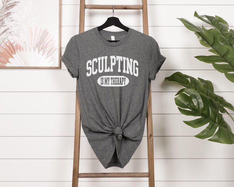 K&ouml;nnte beinhalten: Ein graues T-Shirt mit dem Text "SCULPTING IS MY THERAPY" in wei&szlig;en Blockbuchstaben. Das Shirt ist an der Taille geknotet und h&auml;ngt an einem schwarzen Kleiderb&uuml;gel. Eine Holzleiter und eine gr&uuml;ne Pflanze sind im Hintergrund.