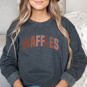 May include: A dark gray sweatshirt with the word "WAFFLES" printed in brown, pink, and orange letters. The sweatshirt has long sleeves and a crew neck.