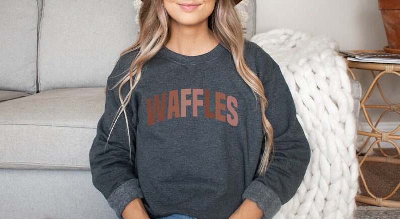 May include: A dark gray sweatshirt with the word "WAFFLES" printed in brown, pink, and orange letters. The sweatshirt has long sleeves and a crew neck.