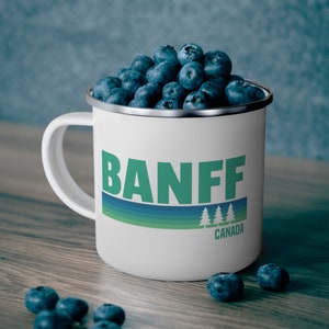 May include: White enamel camping mug with a blue, green, and white stripe design. The mug has the word "BANFF" in green letters and the word "CANADA" in smaller letters below. The mug is filled with blueberries.