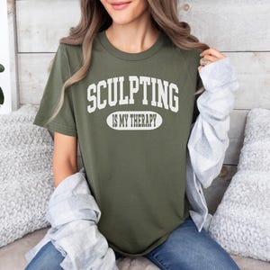 K&ouml;nnte beinhalten: Olivgr&uuml;nes T-Shirt mit dem wei&szlig;en Text "SCULPTING IS MY THERAPY". Der Text ist in gro&szlig;en, geschwungenen Buchstaben &uuml;ber und unter einem wei&szlig;en Oval angeordnet. Das T-Shirt hat einen Rundhalsausschnitt und kurze &Auml;rmel.