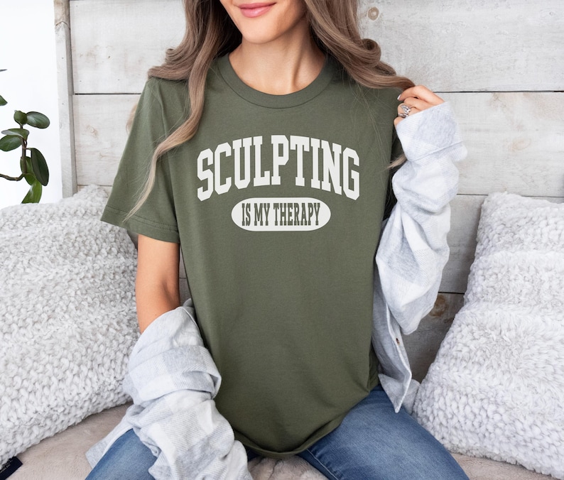 K&ouml;nnte beinhalten: Olivgr&uuml;nes T-Shirt mit dem wei&szlig;en Text "SCULPTING IS MY THERAPY". Der Text ist in gro&szlig;en, geschwungenen Buchstaben &uuml;ber und unter einem wei&szlig;en Oval angeordnet. Das T-Shirt hat einen Rundhalsausschnitt und kurze &Auml;rmel.