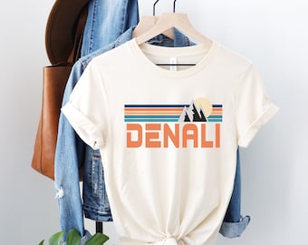 May include: A white t-shirt with a retro-style graphic of mountains and a sun with the word "DENALI" in orange text.