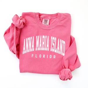 May include: A pink long-sleeved sweatshirt with the text "ANNA MARIA ISLAND FLORIDA" in white block letters. The sweatshirt is folded, with the sleeves cuffed at the wrists. The Comfort Colors tag is visible.
