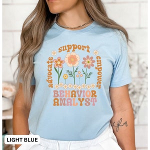 May include: Light blue t-shirt with a floral design and the text "support advocate empower BEHAVIOR ANALYST" in a retro font.