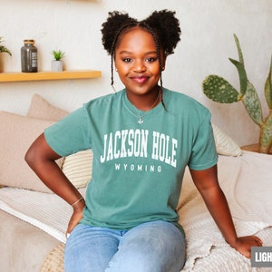 May include: A woman wearing a light green t-shirt with the text "JACKSON HOLE WYOMING" printed on the front. She is sitting on a light beige couch.