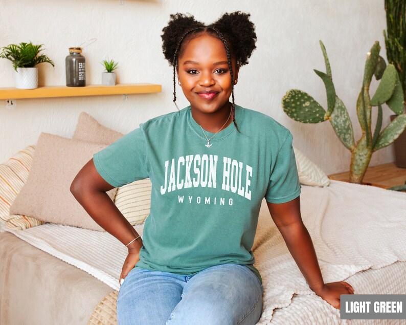 May include: A woman wearing a light green t-shirt with the text "JACKSON HOLE WYOMING" printed on the front. She is sitting on a light beige couch.