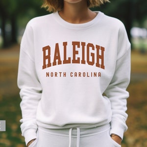 Pode incluir: Camisola branca com um design de arco castanho que diz "Raleigh North Carolina".