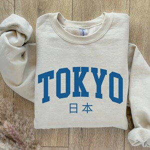 以下が含まれることがあります： 「TOKYO」と「日本」が書かれた、ベージュのトレーナー。フォントは大学風です。