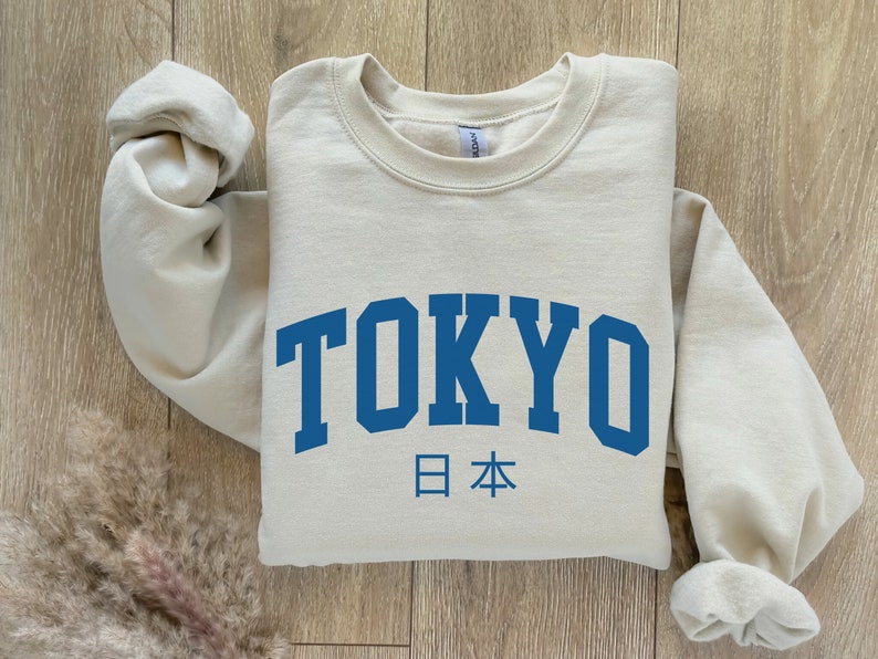 以下が含まれることがあります： 「TOKYO」と「日本」が書かれた、ベージュのトレーナー。フォントは大学風です。