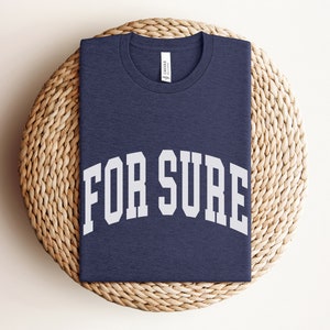 May include: A navy blue t-shirt with the text "FOR SURE" in white, in a collegiate-style font.